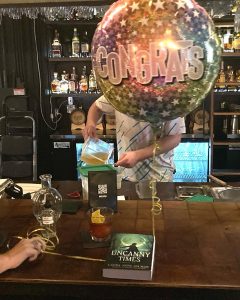 a "congrats!" balloon and a copy of UNCANNY TIMES on a wooden bartop.